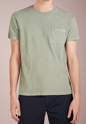 Mann in einem hellgrünen Kurzarm-T-Shirt mit Brusttasche und dunkler Hose, stehend vor einem beigen Hintergrund.