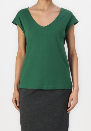 Femme portant un haut vert à manches courtes en col V et une jupe gris foncé, visible du cou jusqu'aux hanches sur un fond uni.