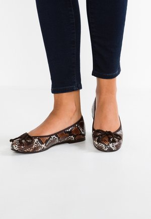 Pieds chaussés de ballerines à motif peau de serpent avec des nœuds à l'avant, portées avec un jean skinny bleu foncé sur un fond blanc.