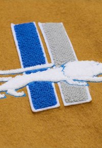 Brauner Stoffhintergrund mit blauen und grauen rechteckigen Flecken; weißes, gesticktes Löwendesign in der Mitte mit strukturierten Akzenten.