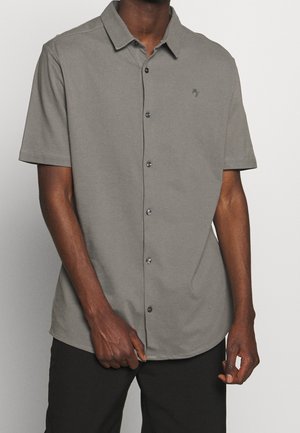 Uomo che indossa una camicia grigia a maniche corte con colletto e un piccolo logo ricamato sul petto, abbinata a pantaloni neri.