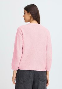 Pulover roz tricotat gros, cu textură cordonată, mâneci largi și un tiv ușor scurtat. Purtați-l cu pantaloni gri închis, cu talie înaltă.