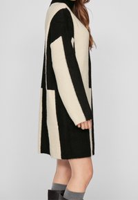 Long cardigan à rayures verticales noires et crème, texture côtelée, devant ouvert et coupe décontractée. Matière douce, silhouette légèrement oversize.