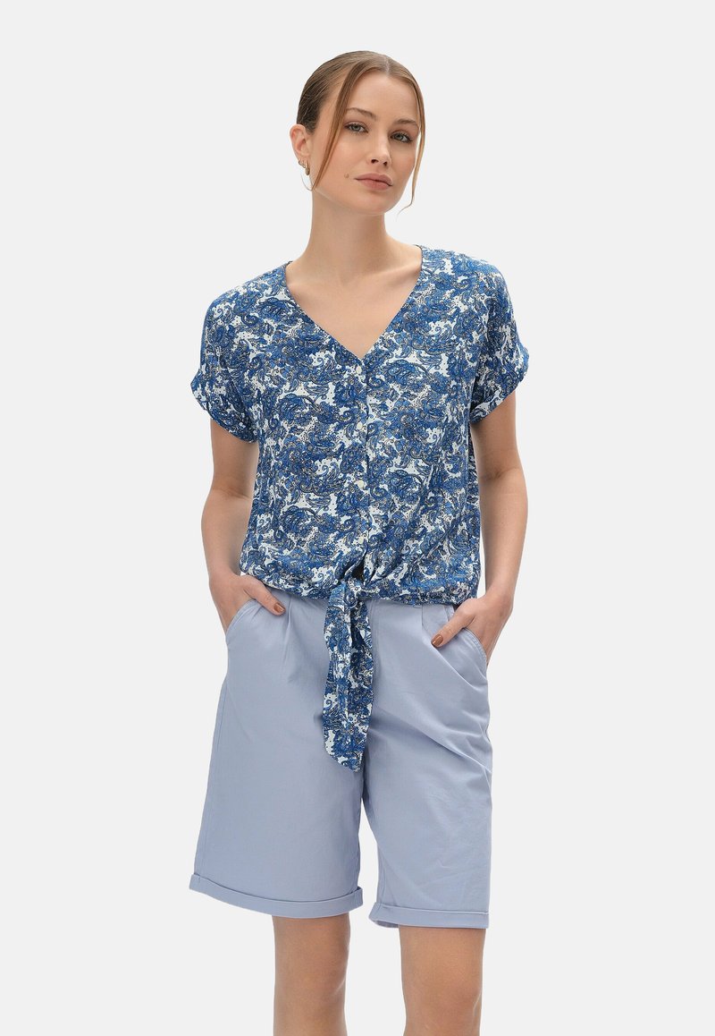 Niebieska bluzka w kwiatowy print z dekoltem w kształcie litery V i węzłem z przodu, zestawiona z jasnoniebieskimi szortami z bocznymi kieszeniami i podwiniętymi mankietami.