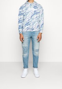 Mikina s kapucí s modro-zeleným vlnkovým vzorem; kombinována se sepranými světle modrými džíny a bílými teniskami.