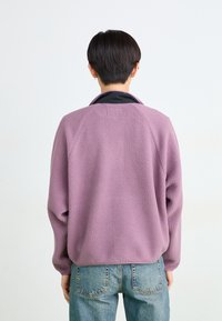 Persona con cabello corto oscuro, vestida con chaqueta de polar morada y jeans azules, vista desde atrás sobre un fondo claro y liso.