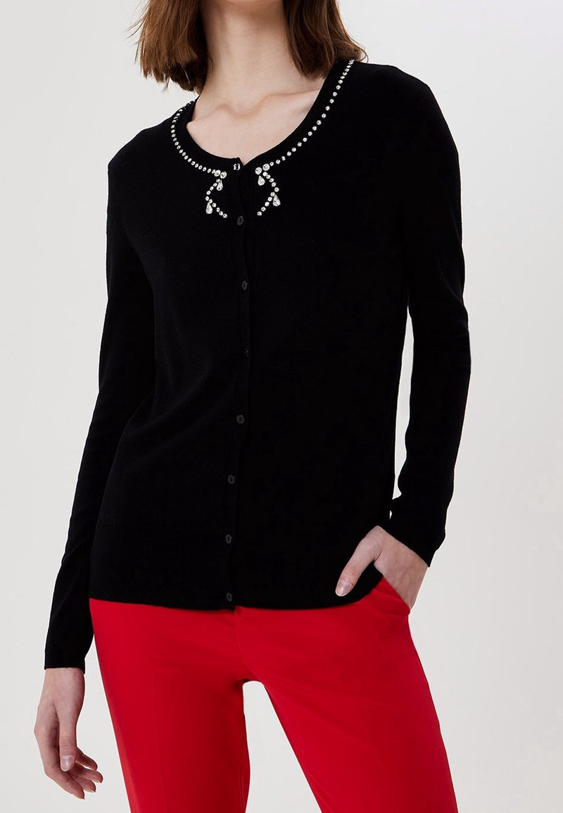 Femme portant un cardigan noir à manches longues boutonné avec des détails en strass au niveau du col et un pantalon rouge, main dans la poche.