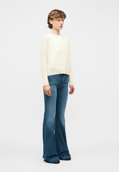 Νεαρός με κοντά μαλλιά φοράει κρεμ πλεκτή ζακέτα με κουμπιά και ψηλόμεσα μπλε καμπάνα τζιν, στέκεται μπροστά σε απλό φόντο.