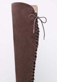 Bota alta de ante marrón con detalle de cordones verticales y un lazo atado en la parte superior sobre un fondo claro y sencillo.