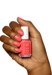 Flacon de vernis à ongles rose corail tenu dans une main marron clair. Les ongles sont vernis dans la même teinte corail. Fini brillant, forme de flacon ronde, capuchon blanc.