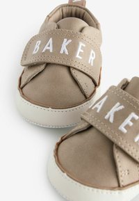 Vaaleat lasten kengät, jotka on valmistettu pehmeästä materiaalista valkoisilla kumipohjilla. Varustettu tarranauhalla, jossa on valkoisella brodeerattu "BAKER".