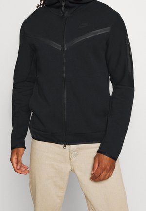 Mann trägt einen schwarzen Kapuzenpullover mit Reißverschluss, diagonalem Streifendetail und Reißverschlusstasche am linken Ärmel, kombiniert mit beigen Hosen.