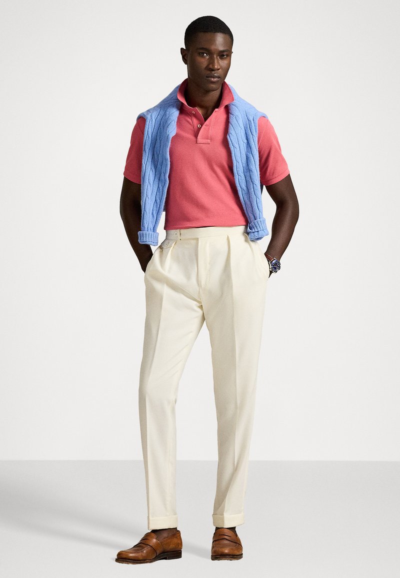 Polo rosa, gilet azzurro chiaro di maglia appoggiato sulle spalle, pantaloni plissettati color crema e scarpe in pelle marrone. Il modello sta in piedi con sicurezza.