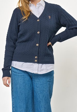 Personne portant un cardigan en maille torsadée bleu marine par-dessus une chemise blanche et un jean bleu, avec une main sur la hanche et l'autre détendue.