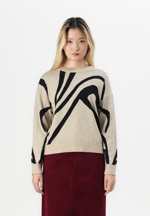 Pull beige avec des motifs abstraits noirs, col rond et manches longues. Associé à une jupe bordeaux, le tissu semble doux et texturé.