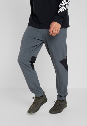 Persona che indossa pantaloni grigi affusolati con toppe nere sulle ginocchia, maglia nera a maniche lunghe e sneaker verdi scuro con lacci.