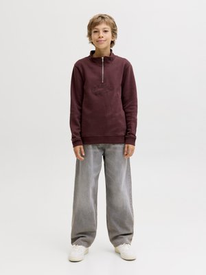 Mladá osoba stojí, oblečená v vínově červené mikině s krátkým zipem, volných šedých džínách a bílých teniskách před jednobarevným světlým pozadím.