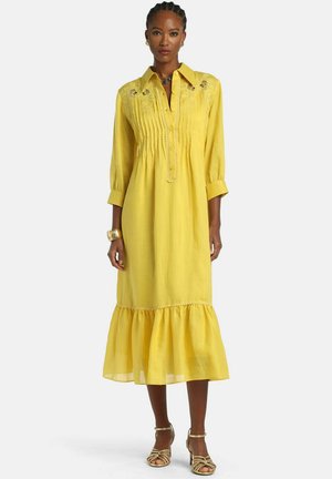 Mujer con un vestido largo amarillo con bordados y pliegues, que presenta un dobladillo con volantes, mangas tres cuartos y sandalias doradas.