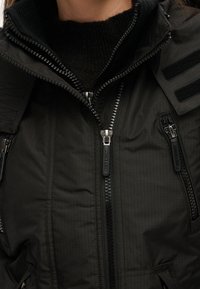 Svart zip-up-jacka med texturerad tyg, flera dragkedjefickor och kardborreaccent på axeln. Har en hög krage för extra värme.