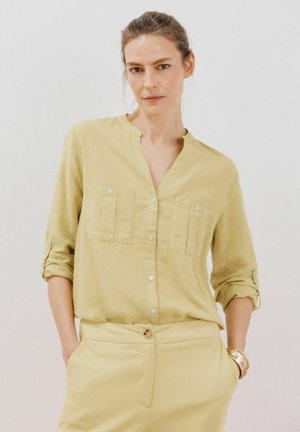 Camicia button-up gialla chiara con due tasche sul petto, maniche arrotolabili e design senza colletto. Tessuto liscio e stile minimalista.