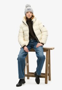 Giubbotto imbottito bianco sporco, dolcevita in maglia marrone, jeans blu e stivali neri con lacci. Berretto in maglia grigia con pompon e dettaglio con toppa.