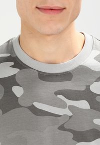 Camiseta de camuflaje gris con cuello redondo y tela texturizada que presenta una variedad de tonos de gris y patrones en blanco.