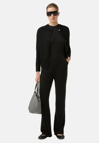 Schwarze Strickjacke mit Knöpfen, eng anliegendes schwarzes Top, maßgeschneiderte schwarze Hose, schwarze Schuhe und eine hellgraue Handtasche. Accessoires umfassen übergroße schwarze Sonnenbrillen.