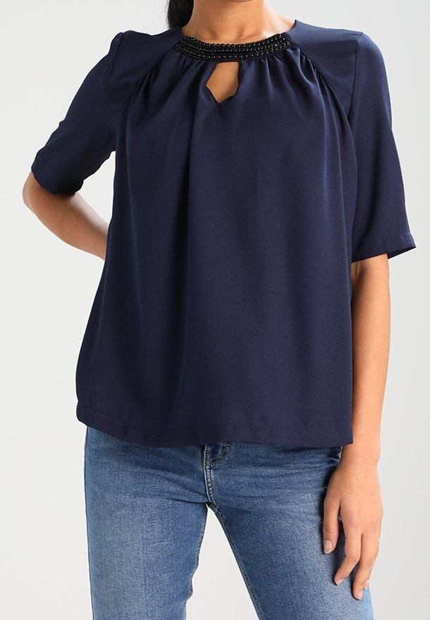 Femme portant un chemisier bleu marine à manches courtes avec un col orné de perles et un détail en forme de goutte, associée à un jean bleu clair, sur fond uni.