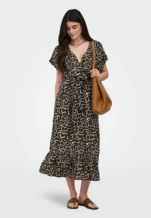 Kvinne iført leopardmønstret midikjole, som holder en brun skulderveske, med langt mørkt hår, gullsmykker og brune flate sandaler.