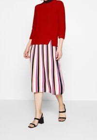 Roter gerippter Pullover mit dreiviertel Ärmeln, kombiniert mit einem Midirock mit vertikalen Streifen in verschiedenen Farben. Schwarze Block-Absatzsandalen.