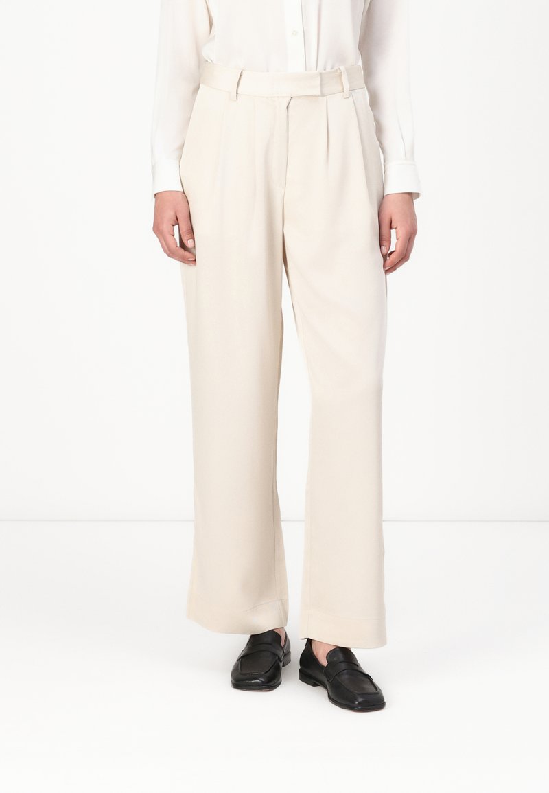Pantalons larges crème avec des plis, confectionnés dans un tissu lisse. Portés avec une chemise blanche et des mocassins noirs, présentant une coupe ajustée.