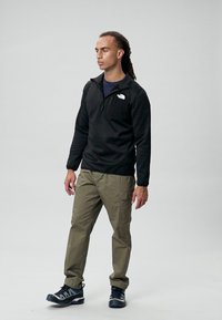 Sudadera de forro polar negra con cuello de cremallera, pantalones cargo olivo y zapatillas deportivas negras con detalles en gris. Hombre de pie contra un fondo neutro.