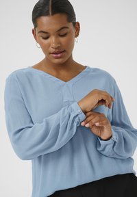 Blouse bleu clair à manches longues avec un col en V, en tissu texturé, avec des poignets boutonnés et une coupe décontractée. Pliages subtils le long des manches.