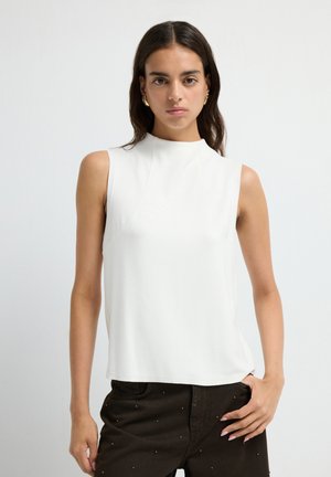 Femme aux cheveux noirs portant un haut sans manches blanc et un pantalon noir avec de petites clous, se tenant contre un fond clair.