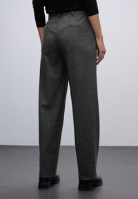 Donkergrijze wijde broek in zachte stof met achterzakken en een gladde tailleband, gedragen met zwarte schoenen tegen een neutrale achtergrond.