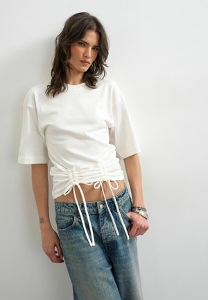 Femme aux cheveux bruns mi-longs, portant un haut blanc froncé à manches courtes et un jean bleu ample sur un fond clair uni.