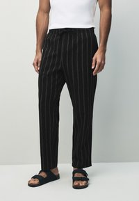 Schwarz gestreifte Hosen mit einem Kordelzug in der Taille, aus einem leichten Stoff gefertigt. Kombiniert mit schwarzen Sandalen mit verstellbaren Riemen.