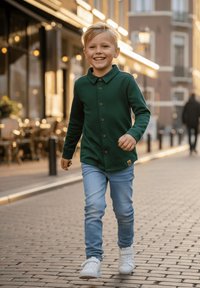 Zielona koszula z długim rękawem zapinana na guziki z kołnierzykiem, połączona z jasnoniebieskimi dżinsami i białymi trampkami, noszona przez chodzącego dziecka na brukowanej ulicy.