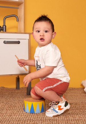 Een kind in een wit T-shirt en rode shorts zit op een blauw en geel drumstel. Grijze kasten en een gestructureerd bruin tapijt zijn op de achtergrond.