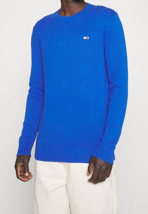 Man wearing bright blue long-sleeve knit sweater with small rectangular logo on chest and off-white pants, standing against plain background.