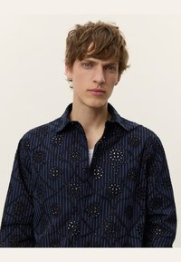 Marineblå skjorte med blomstermønster utskåret design, vertikale hvite striper og krage. Laget av lett stoff med frontåpning med knapper.