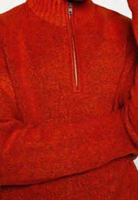 Roter Sweater mit Viertelreißverschluss, gefertigt aus strukturiertem, wollähnlichem Stoff. Verfügt über einen hohen Kragen und ein dezentes, gesprenkeltes Muster im gesamten Material.