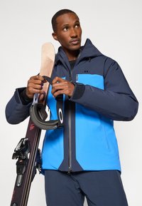 Kék és sötétkék sídzseki, cipzárral elöl, kapucnival és tépőzáras mandzsettákkal. A személy síléceket és egy fekete pántot tart, piros részletekkel.