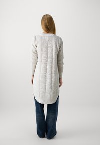 Langer, hellgrauer Strickcardigan mit Zopfmuster, gerippten Bündchen und einem leicht verlängerten Rücken. Getragen über dunkelblauen weit geschnittenen Jeans.