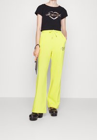 Svart t-shirt med metallisk "LOVE MOSCHINO" design, kombinerad med neongula byxor med vida ben som har en logotyp. Svarta plattformsklackar med guldaccenter.