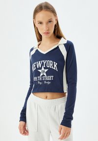 Top corto navy de manga larga, con un cuello blanco y acentos laterales en contraste. El texto impreso incluye "NEW YORK 89.TH STREET" y una estrella.