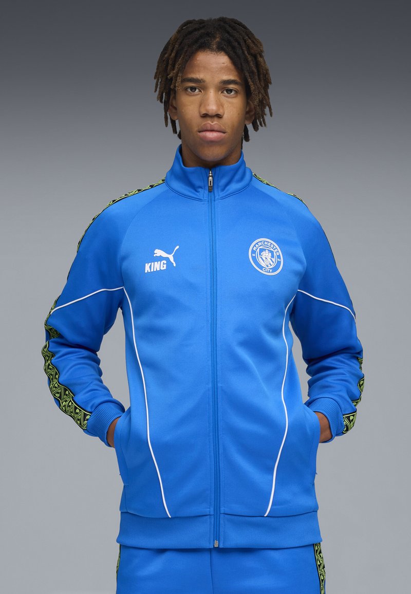 Sininen vetoketjullinen urheilu takki, jossa on valkoisia yksityiskohtia, vihreät kuviolliset sivuhaitarit ja Manchester Cityn logo rintaan ommeltuna.