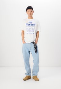 T-shirt blanche avec un motif "Red Bull Racing" bleu, jean bleu clair, bandana noir dans la poche et baskets beiges. Tenue simple et décontractée.