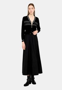Cardigan nero con zip e dettagli a strisce grigie, indossato sopra una gonna nera. Stivali neri con tacco e dettagli borchiati completano l'outfit.