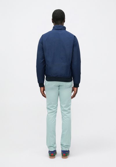 Navy bomber jacket with high collar, ribbed cuffs, worn with light green pants and multicolored shoes, viewed from the back.